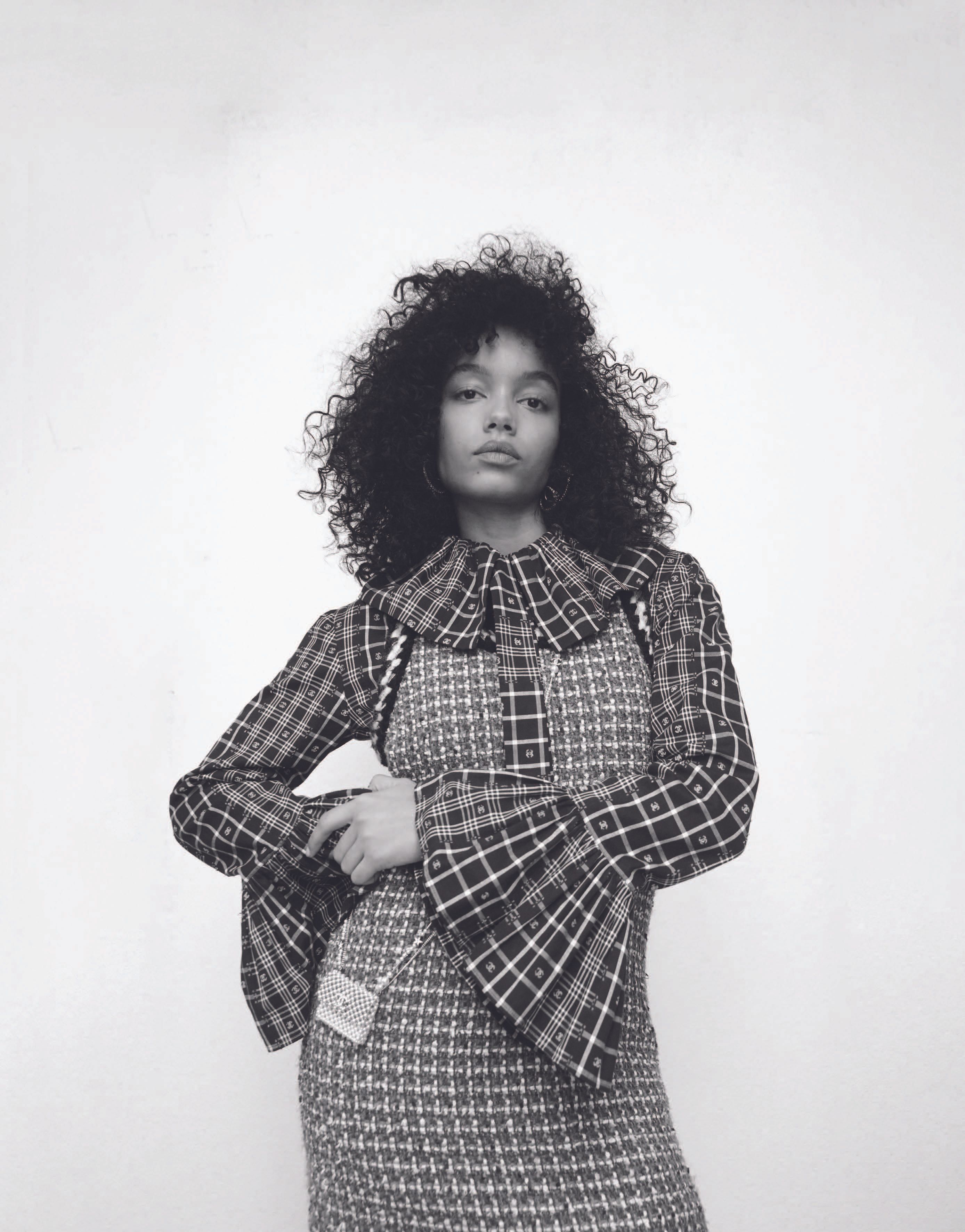 Monochrome portrait of a young woman with curly hair wearing a plaid dress with a ruffled collar, standing against a white wall.