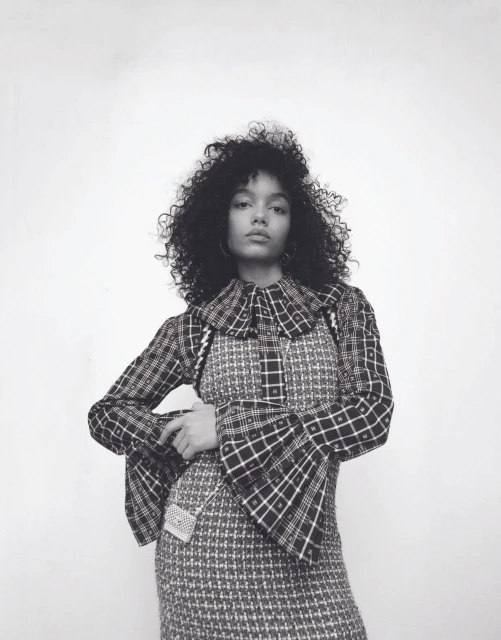 Monochrome portrait of a young woman with curly hair wearing a plaid dress with a ruffled collar, standing against a white wall.