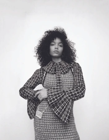Monochrome portrait of a young woman with curly hair wearing a plaid dress with a ruffled collar, standing against a white wall.