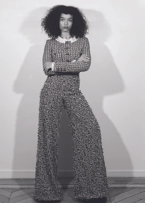 Whitney Peak in a patterned tweed jacket and trousers posing against a plain wall with a sharp shadow cast behind her.