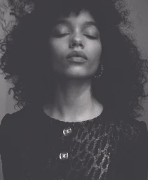 Portrait of Whitney Peak with eyes closed, wearing a dark textured top with metallic buttons and hoop earrings against a dark background.