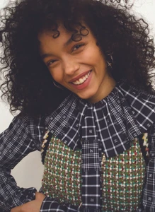 Portrait of Whitney Peak with dark curly hair wearing a plaid blouse and textured vest against a plain white background