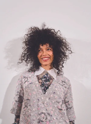 Portrait of Whitney Peak with voluminous curly hair wearing a textured patterned top against a plain white background.