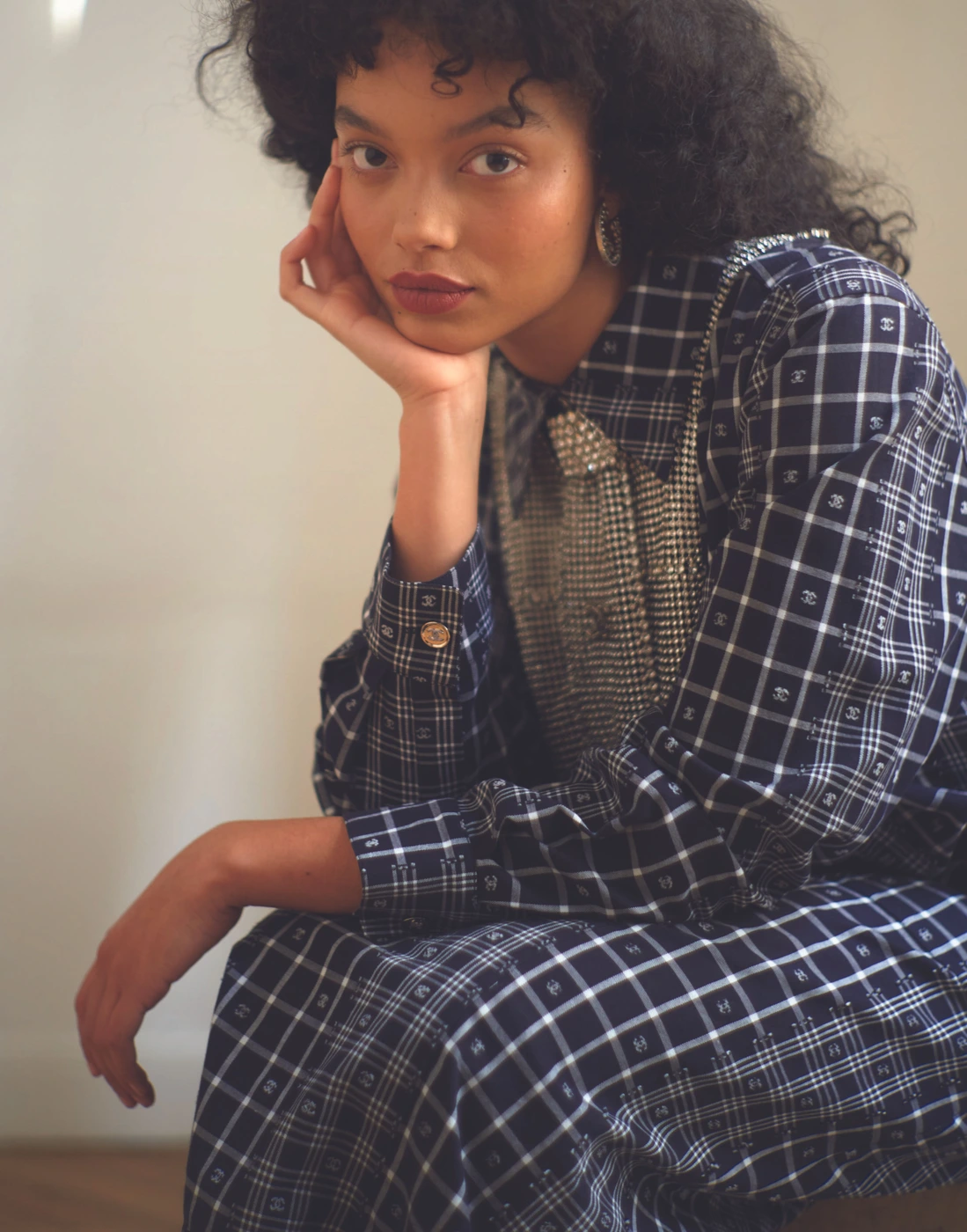 Whitney Peak wearing a blue plaid collared shirt and skirt with her chin resting in her hand against a neutral background.