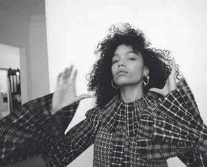 Monochrome portrait of Whitney Peak with dark curly hair wearing a plaid ruffled top and hoop earrings against a white wall.