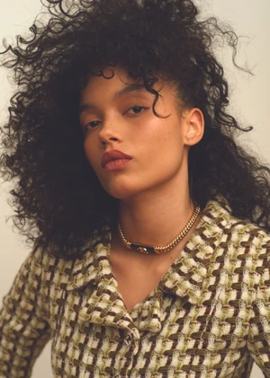 Portrait of Whitney Peak wearing a patterned tweed jacket and gold chain necklace against a light background