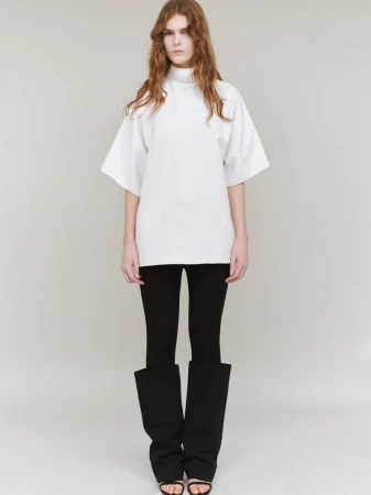 Model standing against a plain grey background wearing a white tunic, black trousers, and tall black boots.