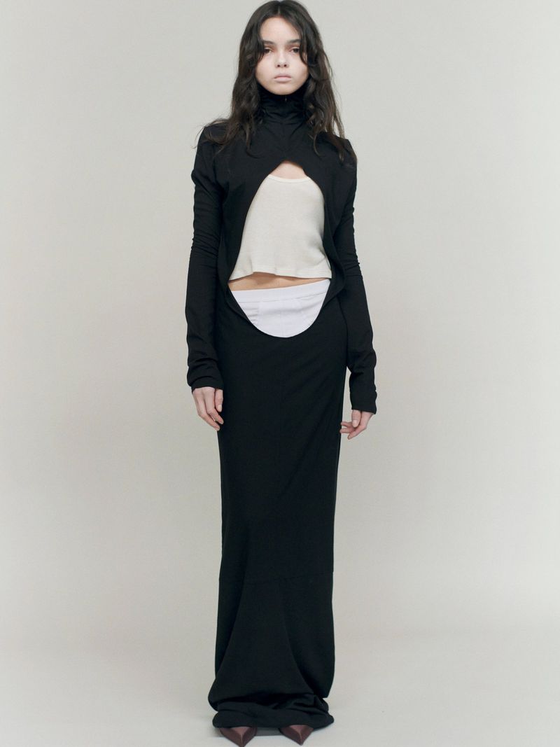 Full-length studio shot of a woman in a black long-sleeved cut-out dress, neutral top, and heels against a light background. Fashion editorial.