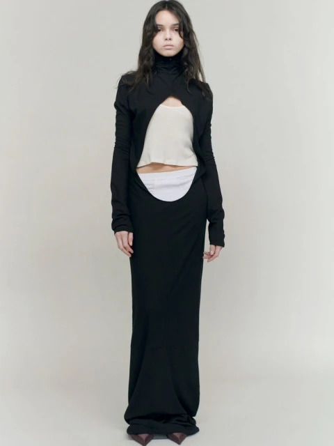 Full-length studio shot of a woman in a black long-sleeved cut-out dress, neutral top, and heels against a light background. Fashion editorial.