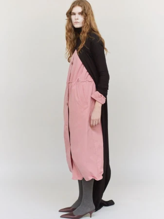 Model standing against a plain white background wearing a pink coat layered over a black top and dark grey stockings.