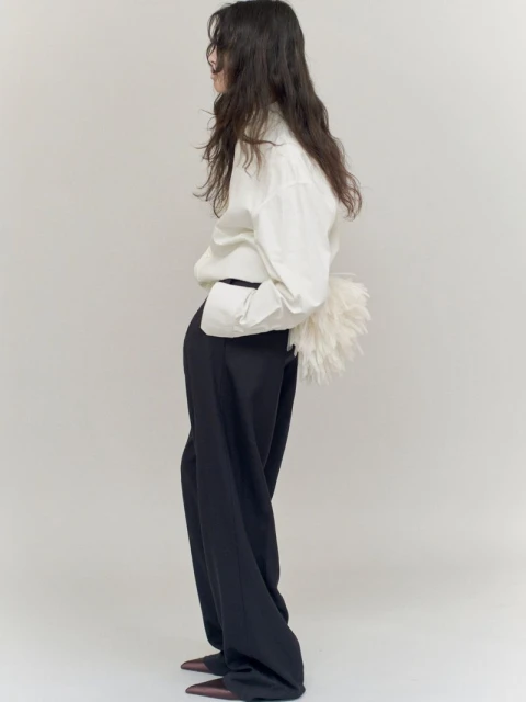 Fashionable woman in a white button-down shirt and flowing black pants stands in a photo studio with a fluffy purse.
