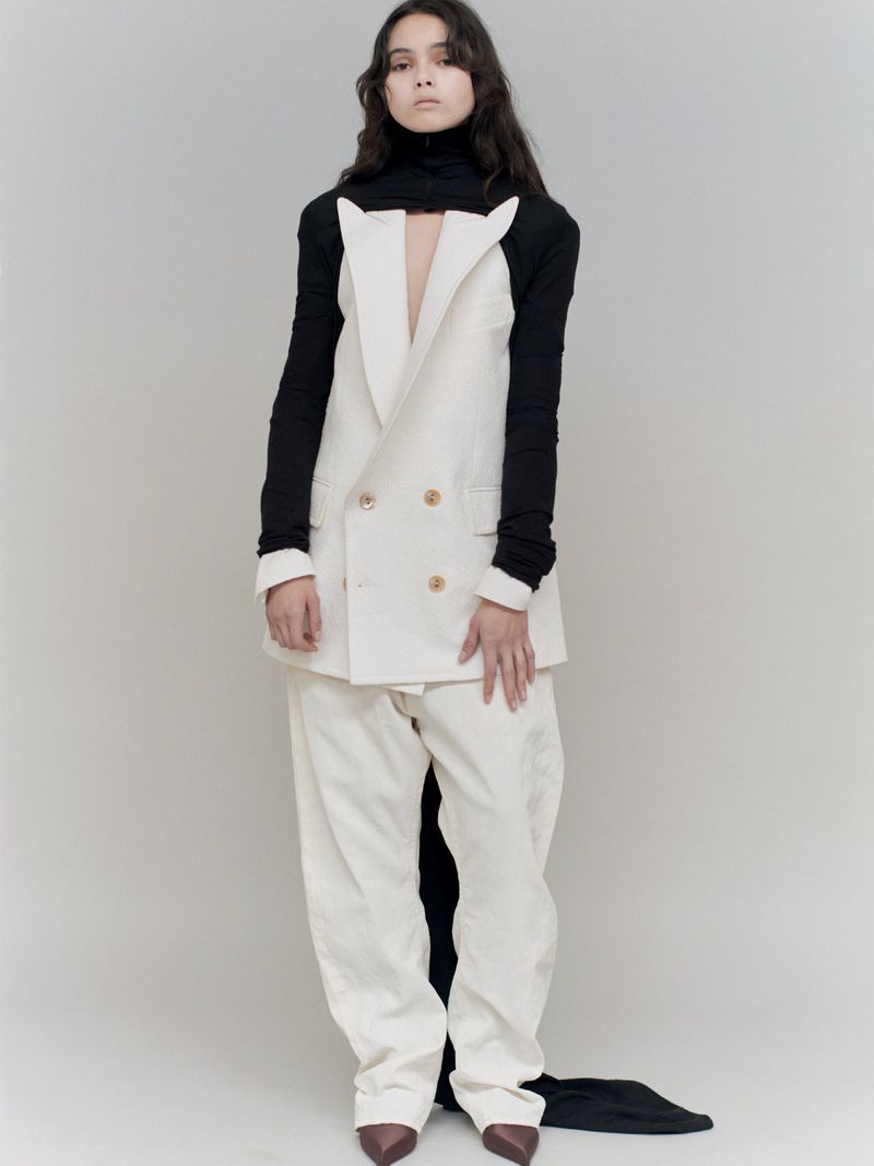 A fashion portrait of a woman in a double-breasted white blazer, matching pants, black turtleneck, and brown heels against a neutral backdrop.