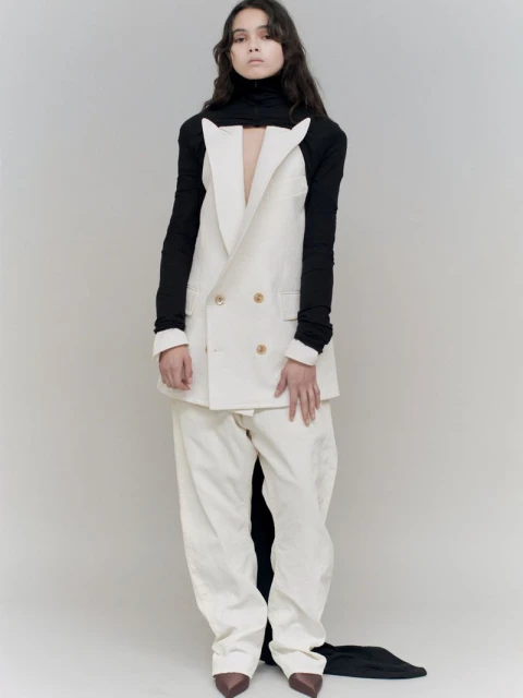 A fashion portrait of a woman in a double-breasted white blazer, matching pants, black turtleneck, and brown heels against a neutral backdrop.