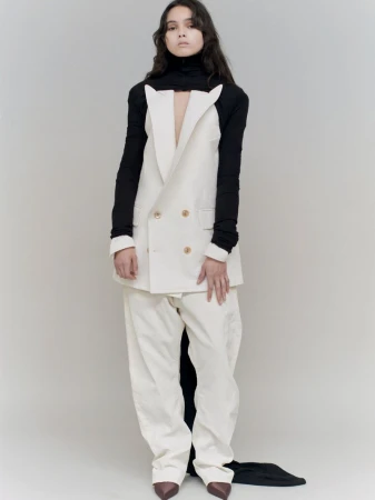 A fashion portrait of a woman in a double-breasted white blazer, matching pants, black turtleneck, and brown heels against a neutral backdrop.
