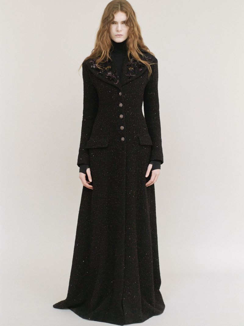 Model wearing a long black coat with embellished lapels standing against a neutral background.