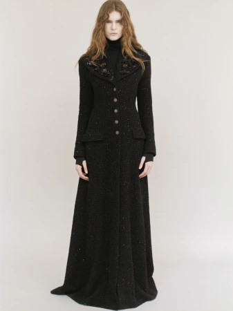 Model wearing a long black coat with embellished lapels standing against a neutral background.