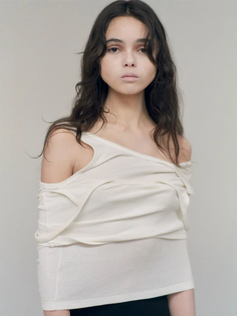 A young adult woman poses in a studio with soft lighting, wearing a draped off-the-shoulder top and dark bottoms against a neutral backdrop.