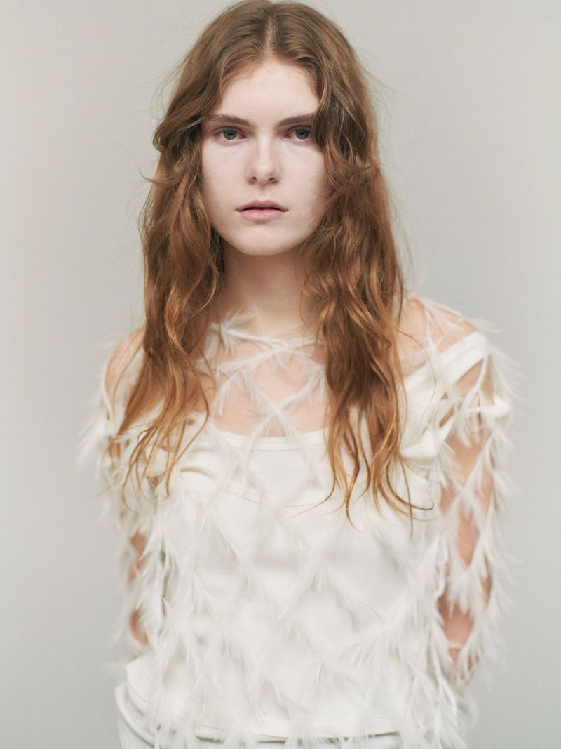 Portrait of a woman with long auburn hair wearing a white top with feather details against a neutral backdrop in an indoor setting.
