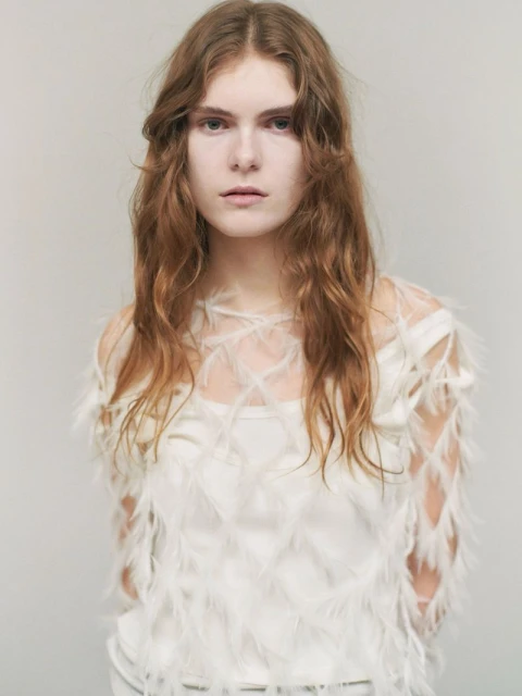 Portrait of a woman with long auburn hair wearing a white top with feather details against a neutral backdrop in an indoor setting.