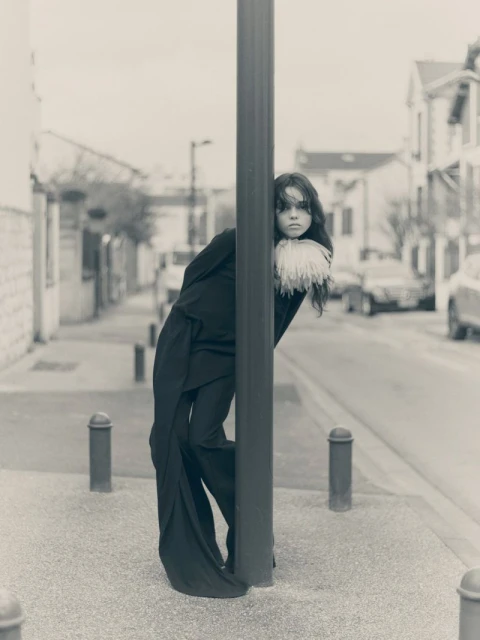 Monochrome portrait of a young woman in a dark suit with white feathered collar, leaning against a pole in an urban street setting.