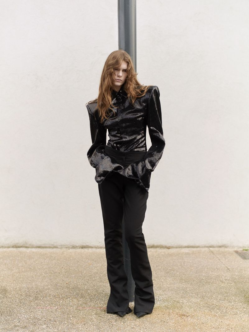 Model poses in a black shiny top and black pants in an outdoor setting with a neutral background, showcasing fashion and style.