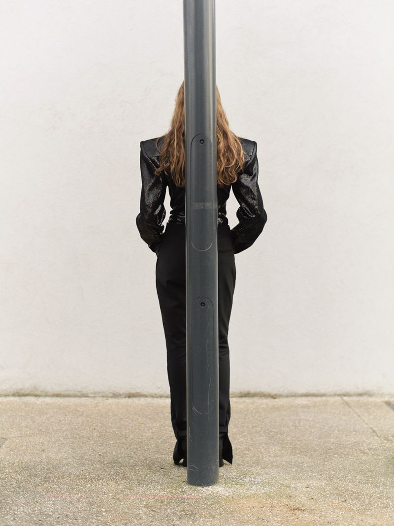 Back view of an adult female in a dark, glittering jacket and black pants, standing behind a grey metal pole outdoors on concrete.