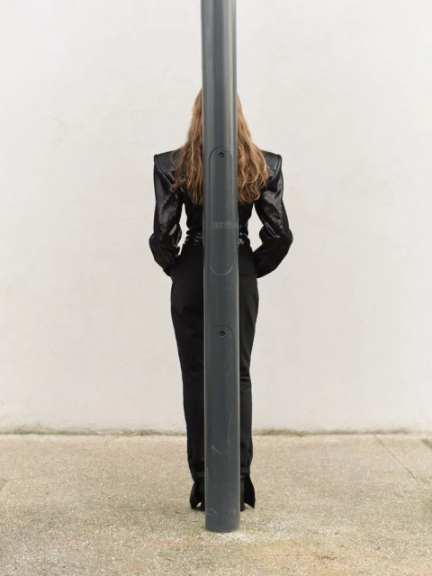 Back view of an adult female in a dark, glittering jacket and black pants, standing behind a grey metal pole outdoors on concrete.