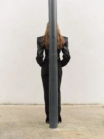 Back view of an adult female in a dark, glittering jacket and black pants, standing behind a grey metal pole outdoors on concrete.