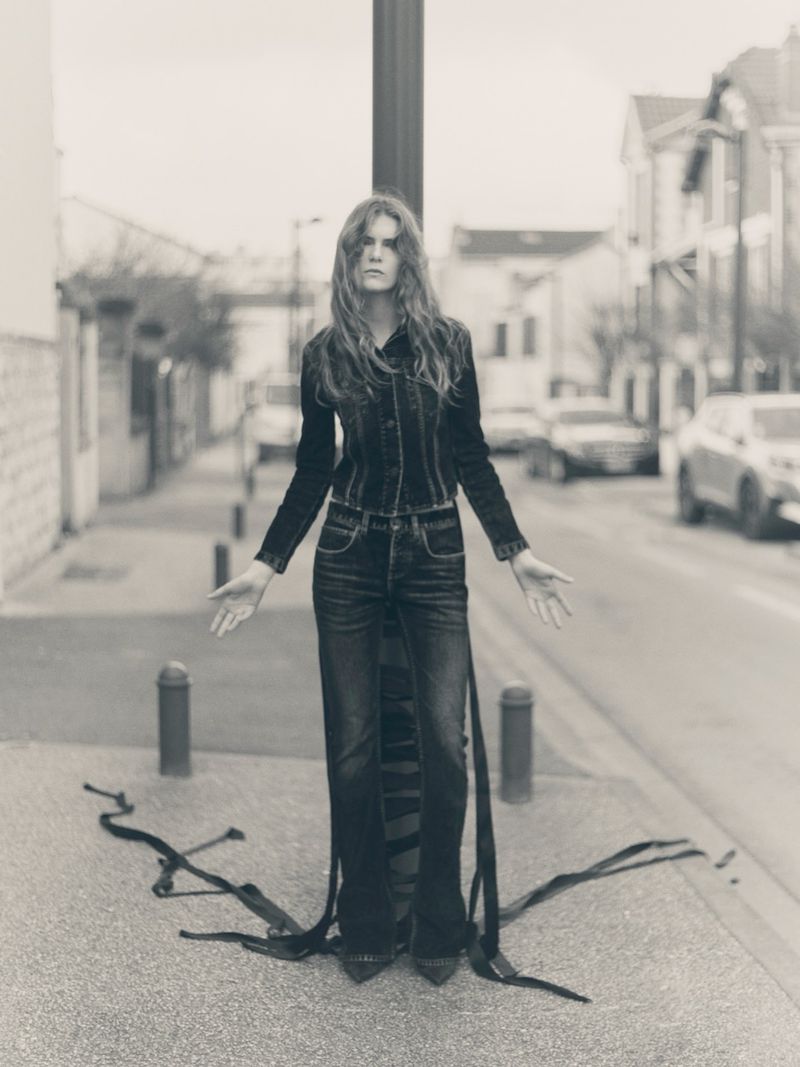 A woman in a denim outfit stands on a city street with ribbons flowing from her legs, in a black and white fashion-style photo.