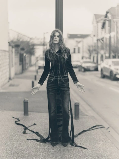 A woman in a denim outfit stands on a city street with ribbons flowing from her legs, in a black and white fashion-style photo.