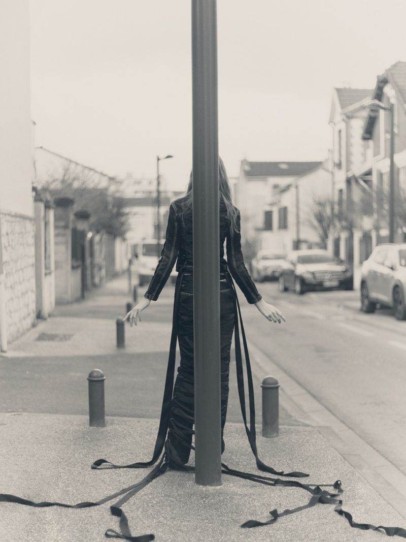 Back view of a slender woman wearing a dark dress with long ribbons standing on a city street next to a pole in monochrome.