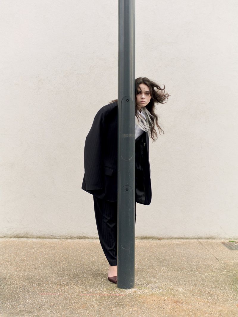 A young woman with long dark hair poses in a black suit, partially concealed by a gray pole in an outdoor urban setting.