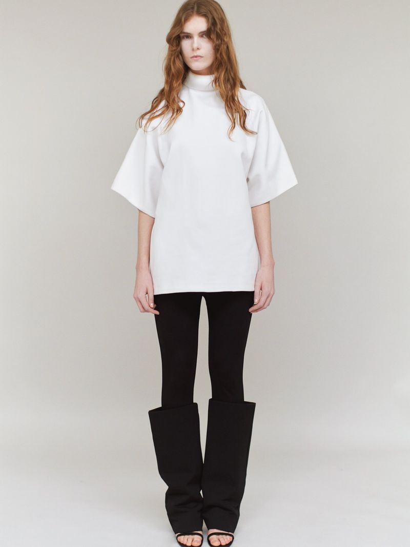Model standing against a plain grey background wearing a white tunic, black trousers, and tall black boots.