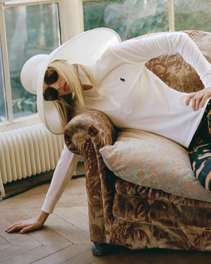 Blonde model wearing a white polo shirt, dark sunglasses, and an oversized white hat reclining on a patterned sofa