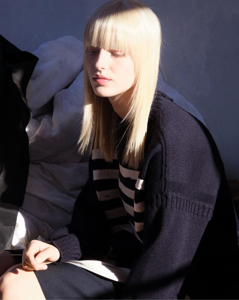 Model with long blonde hair and bangs wearing a dark sweater against an abstract background