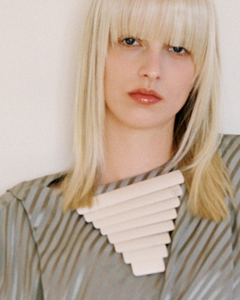Portrait of a blonde woman wearing a striped top and a sculptural necklace against a plain wall.