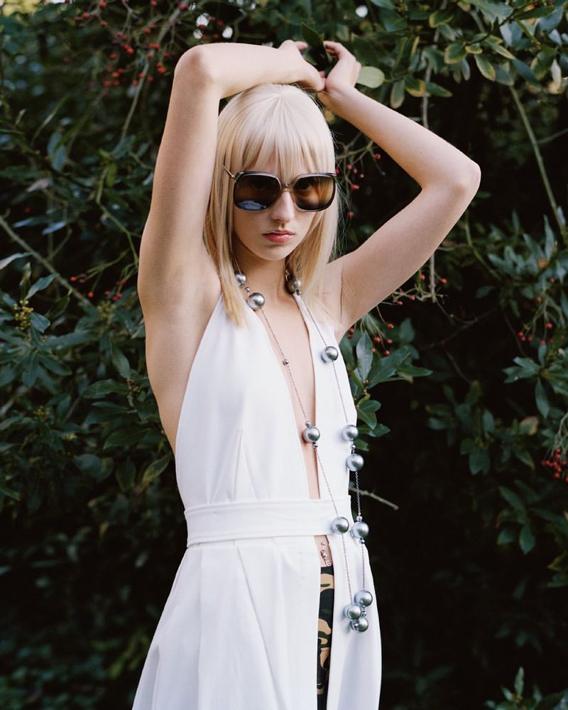 Model with blonde hair wearing oversized sunglasses, a white halter dress, and a long beaded necklace outdoors in front of foliage