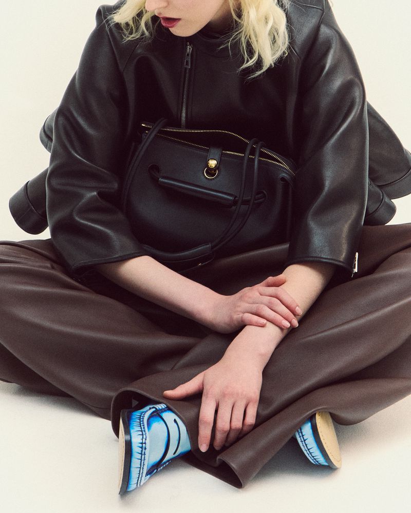 Fashion shot of a person wearing a black leather jacket, brown leather pants, and blue shoes, seated against a white background.
