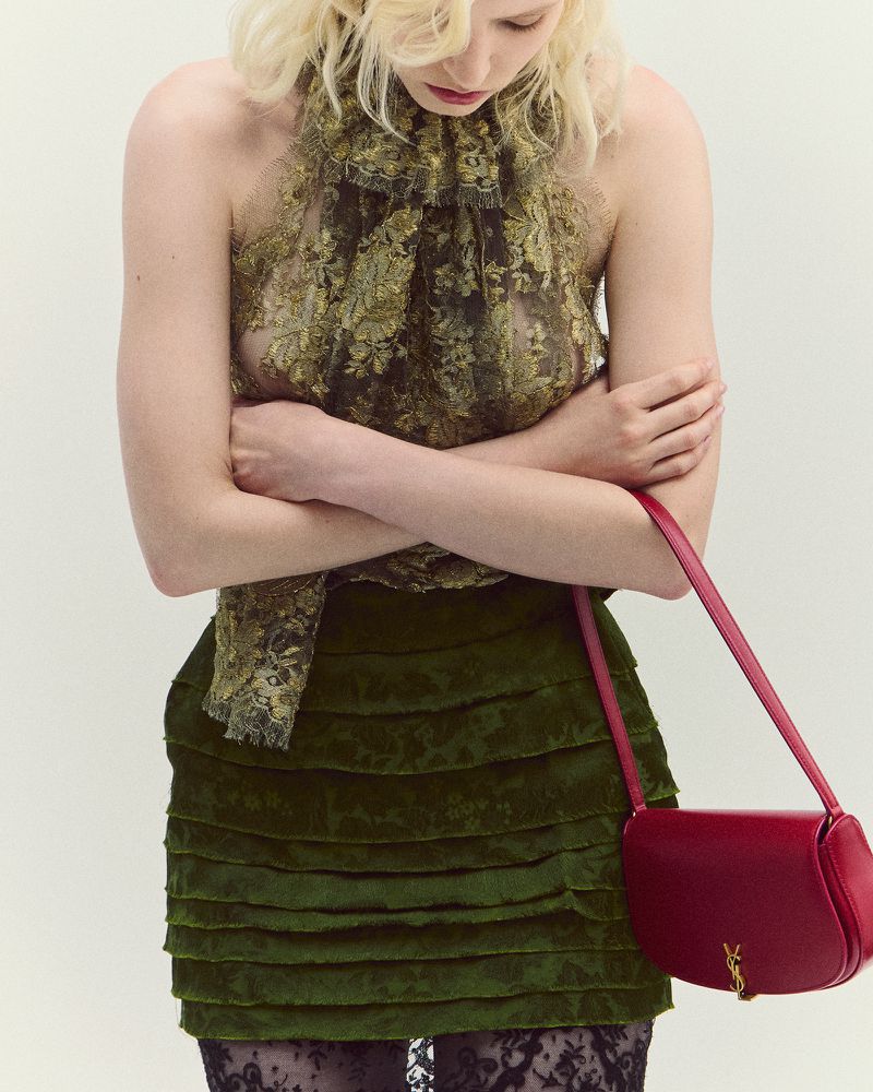 A woman with fair hair wears a floral top and layered skirt, accessorized with a red leather purse and black lace tights against a plain backdrop.