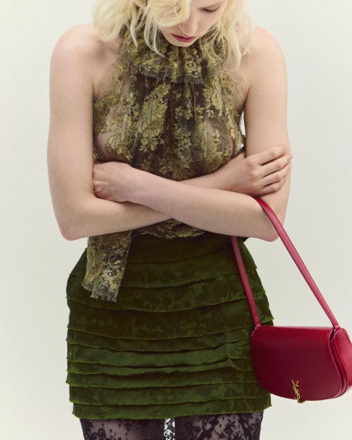 A woman with fair hair wears a floral top and layered skirt, accessorized with a red leather purse and black lace tights against a plain backdrop.