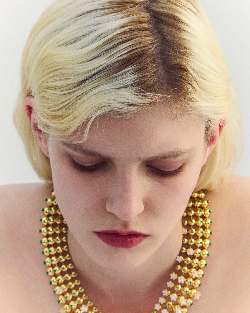 Close-up portrait of a fair-skinned woman with blonde hair looking down while wearing a beaded gold and green necklace against a white background.