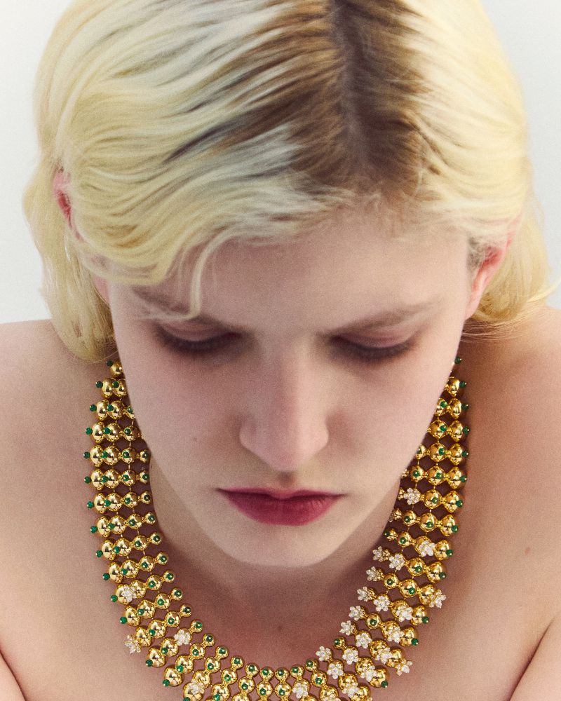 Close-up portrait of a blonde model wearing an elaborate gold and green beaded statement necklace against a plain white background.