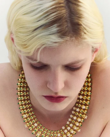 Close-up portrait of a blonde model wearing an elaborate gold and green beaded statement necklace against a plain white background.