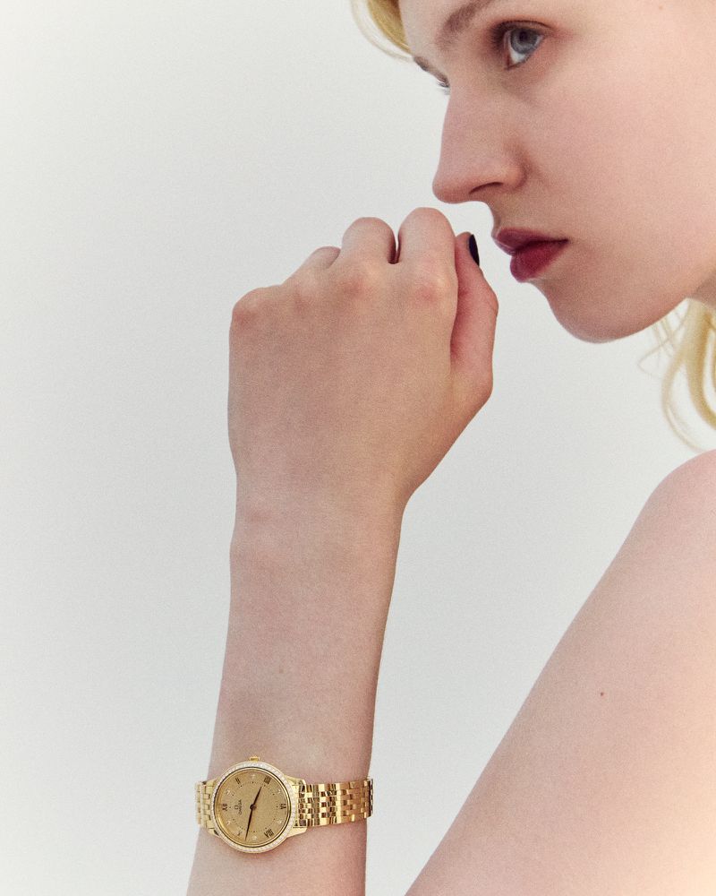 Close-up shot of a young woman with fair skin and blonde hair, showcasing a gold watch on her wrist against a plain white background.