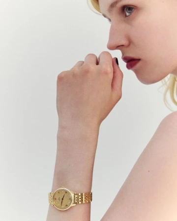 Close-up shot of a young woman with fair skin and blonde hair, showcasing a gold watch on her wrist against a plain white background.
