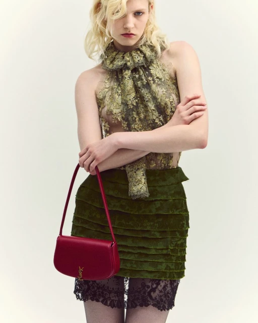A fair-skinned model showcases a Saint Laurent shoulder bag, posing in a lace top and tiered green velvet skirt against a light background.