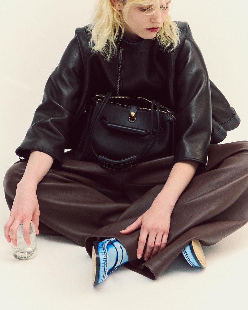 A fair-skinned woman in a black leather jacket and brown pants sits cross-legged holding a glass, a black handbag is next to her.