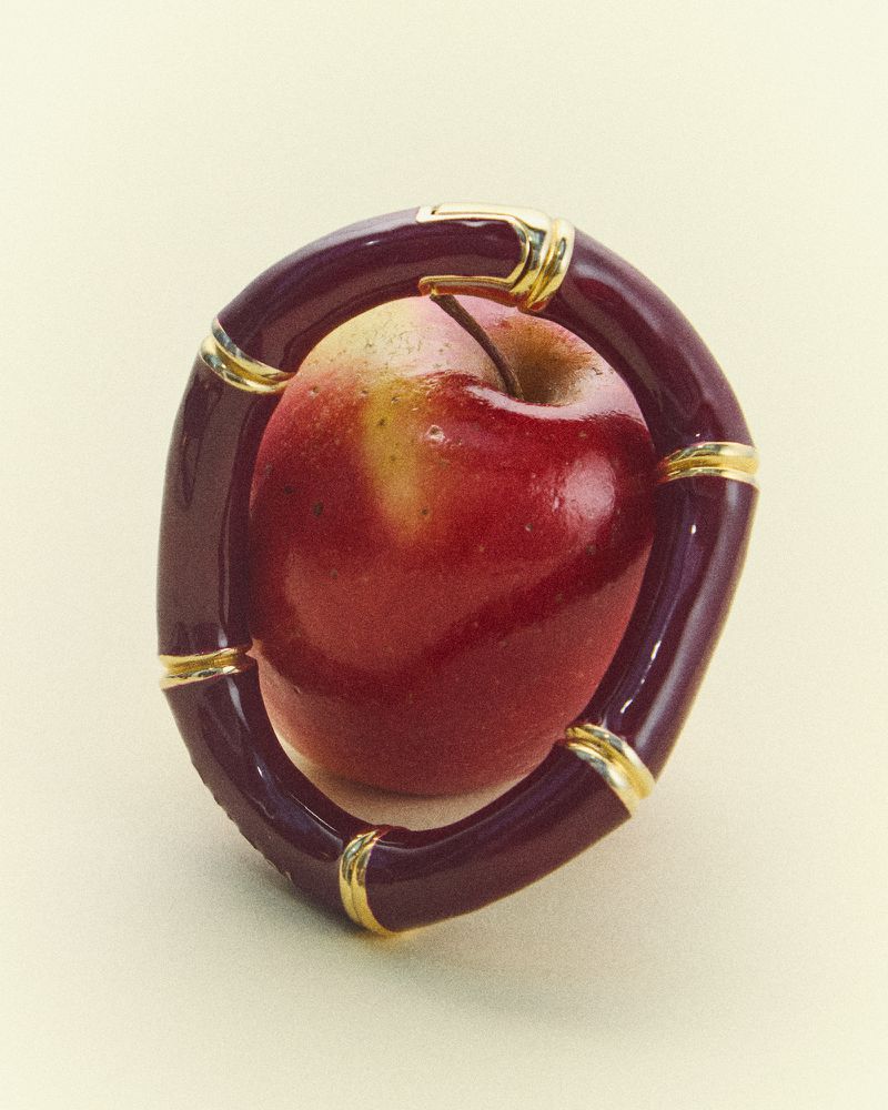 Close-up of a red apple nestled within a dark burgundy bracelet featuring gold accents, set against a soft, creamy backdrop.