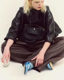 Model with blonde hair wearing a black leather top and brown trousers holding a glass against a white background