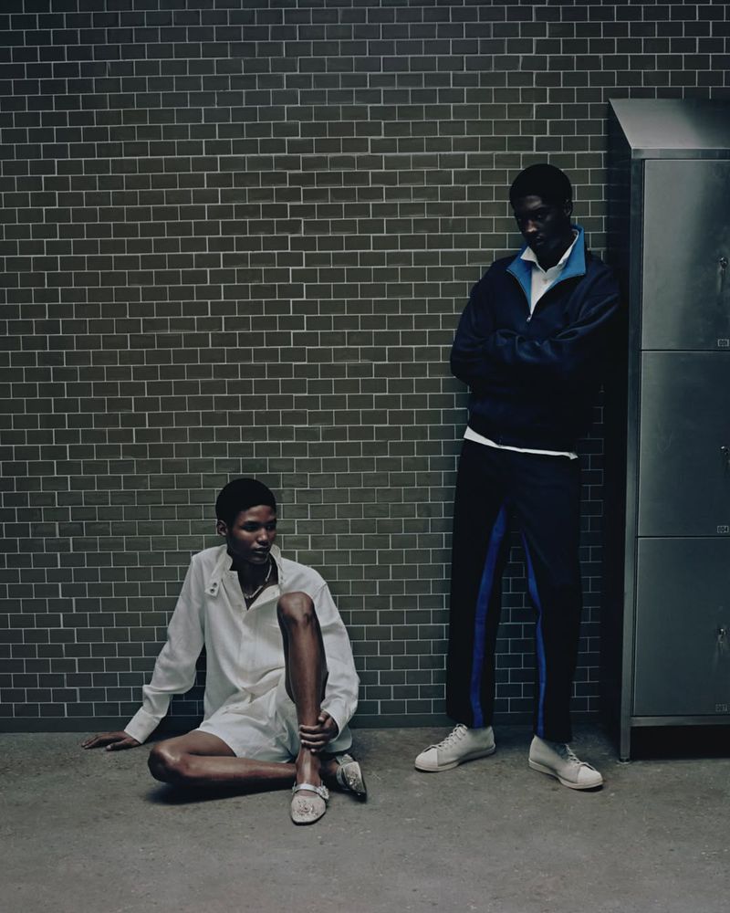 Two Black men pose in athletic wear against a brick wall and metal lockers, one sitting on the floor, the other standing.